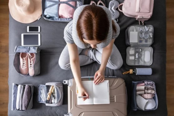 Top view of a woman packing travel essentials including shoes, toiletries, and electronics on a black mat, preparing for an international trip.