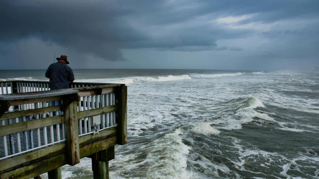 An elderly man stands on a wooden pier overlooking the stormy ocean, symbolizing peaceful retirement moments in Panama amidst dramatic weather.