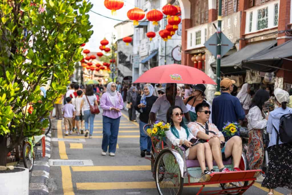 An image of a busy street in Malaysia featuring tourists and locals, with traditional red lanterns hanging overhead, showcasing the vibrant culture and lively atmosphere of the area.