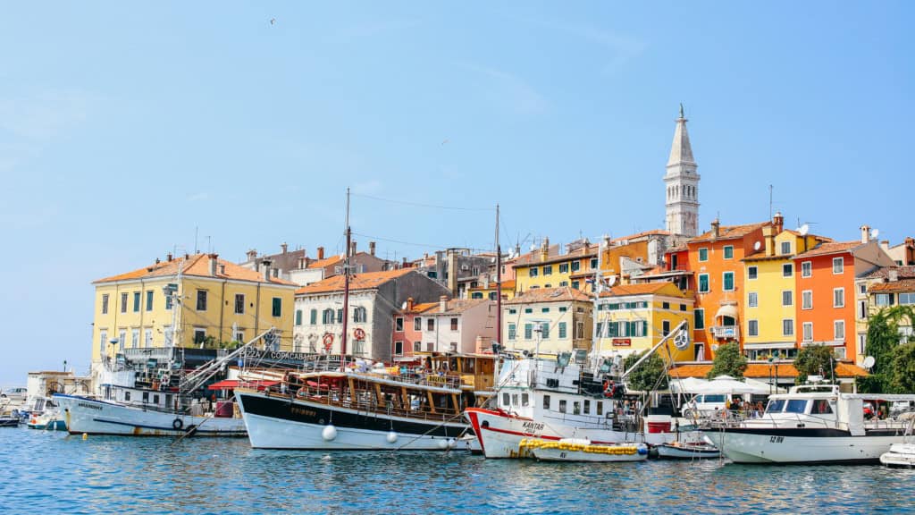 A scenic view of a marina filled with boats and yachts, with vibrant yellow, orange, and pink buildings in the background, set against a clear blue sky in a picturesque seaside town.
