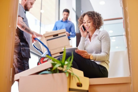 Woman talking on the phone while sitting on a box, with two men in the background handling packages in a modern office setting.