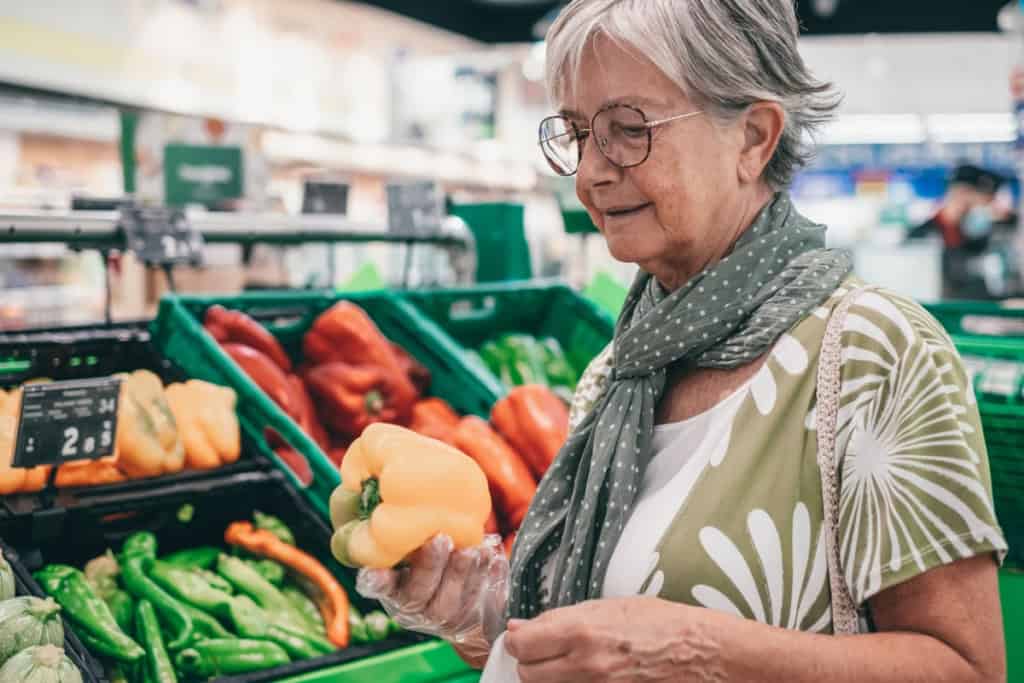 Senior woman shopping for fresh bell peppers at a grocery store, selecting colorful vegetables in the produce section, showing how tackling culture shock and daily life abroad is an important part of retirement planning for expats
