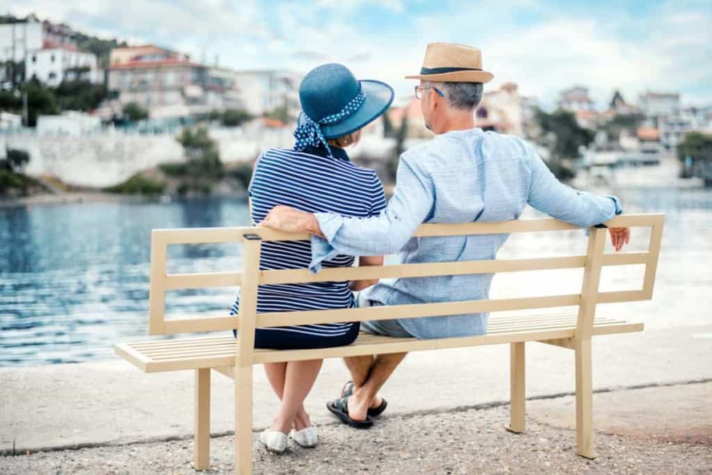 Elderly couple sitting on a bench by the water, enjoying a scenic view, representing international insurance options for expats and retirees.