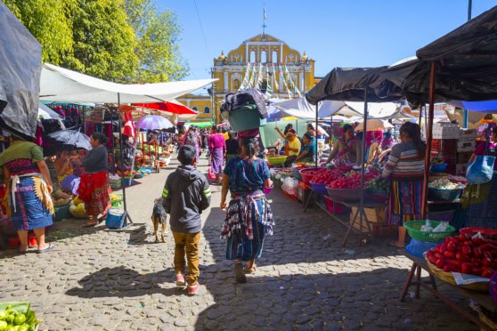 A lively outdoor market bustling with vendors and visitors, featuring vibrant displays of fresh produce, textiles, and local goods under bright sunlight, with a historic building in the background.