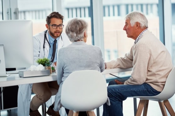 Medical consultation between a doctor and elderly couple in an office setting, discussing health insurance options for international citizens.