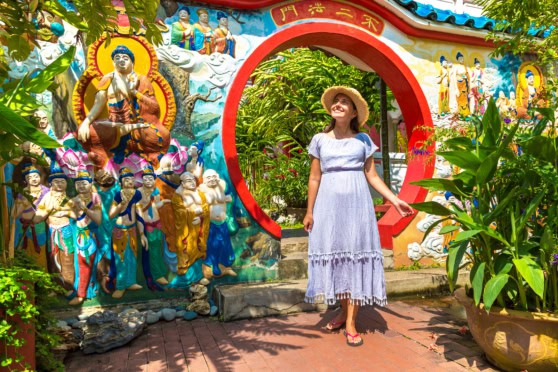 Woman in a white dress and straw hat standing near colorful Asian temple wall with statues and lush greenery.