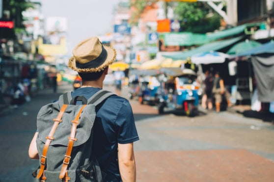 An expat or tourist walking through a busy street in Thailand, carrying a backpack and wearing a hat, with vibrant market stalls and city life in the background.