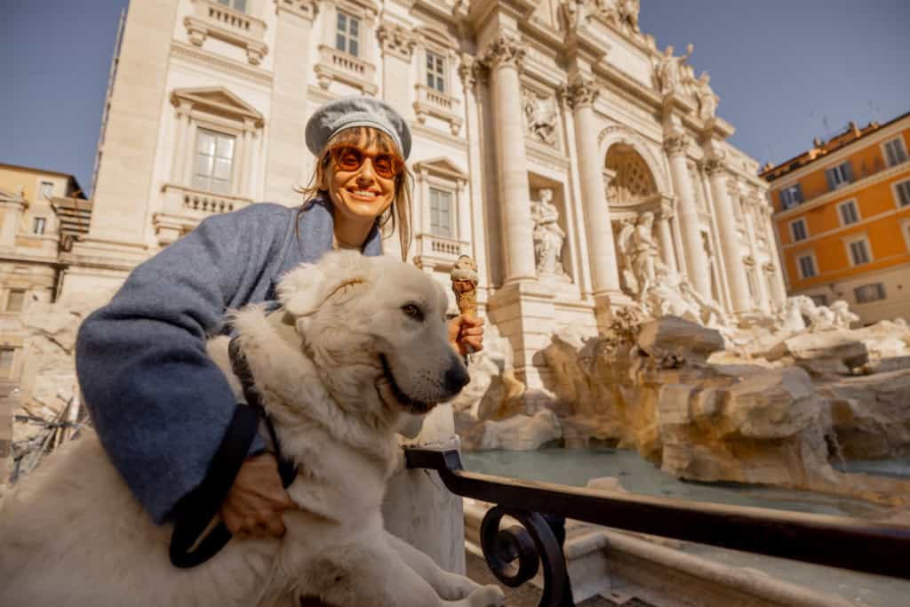 A cheerful woman wearing sunglasses and a gray coat with her large white dog in front of a historic European fountain and ornate building.