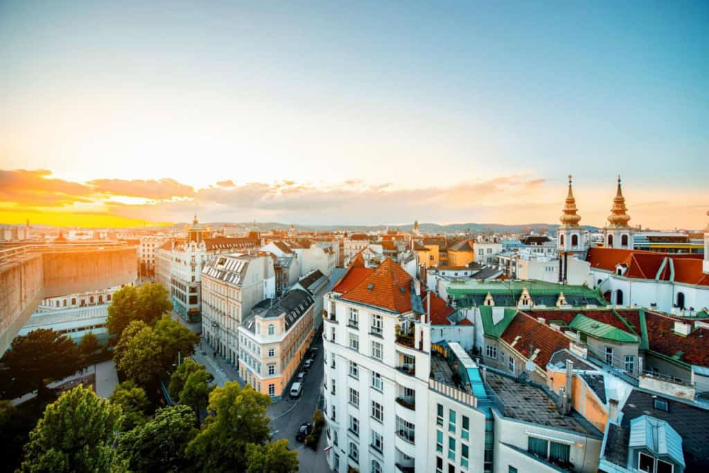 Urban cityscape featuring historic European architecture with colorful rooftops and church spires during sunset.