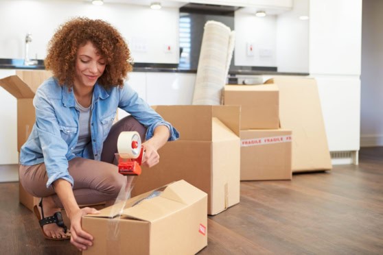 Woman packing a cardboard box with packing tape in a modern apartment, preparing for international relocation or travel.