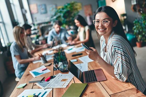 Diverse group of professionals collaborating in a modern office, with a smiling woman in the foreground holding a smartphone, representing international insurance services.