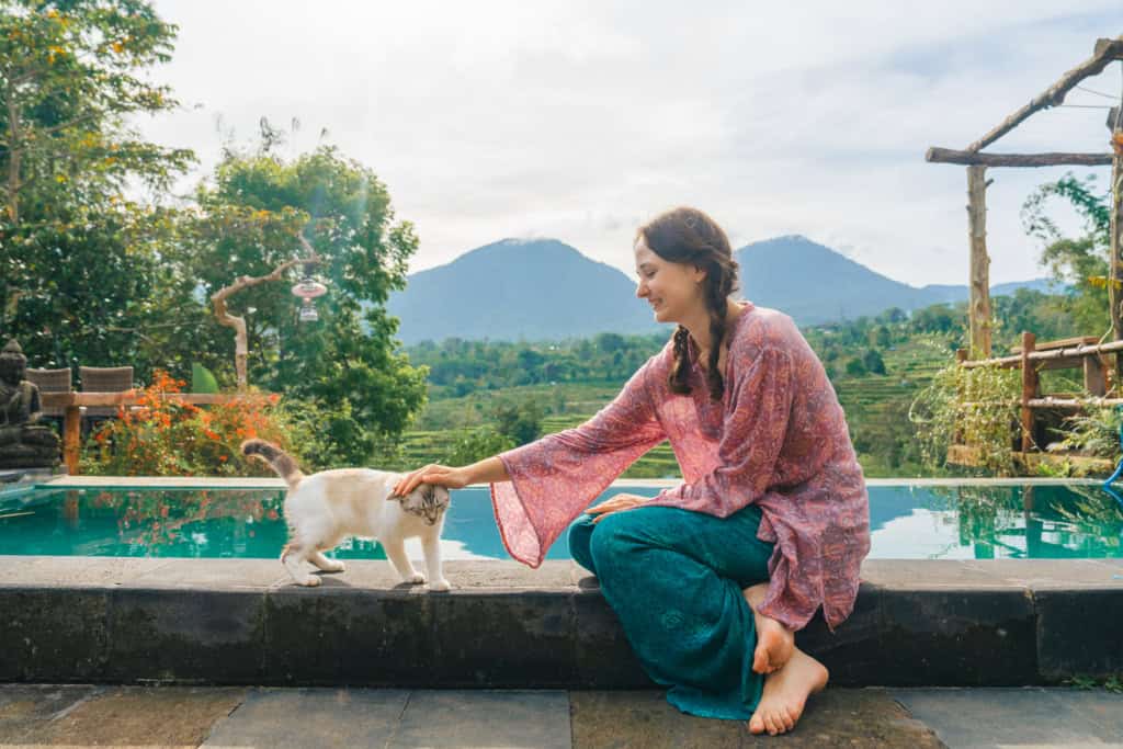 An expat woman in a pink patterned blouse and blue jeans kneels by a pool, gently touching a playful kitten. The background features lush greenery, mountains, and a partly cloudy sky, creating a peaceful, na.