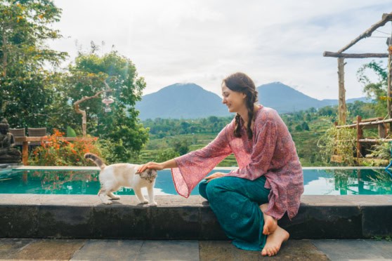 A woman in a pink patterned blouse and blue jeans kneels by a pool, gently touching a playful kitten. The background features lush greenery, mountains, and a partly cloudy sky, creating a peaceful, na.