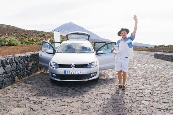 A cheerful man in a hat and casual clothing waves next to a parked Volkswagen car on a volcanic landscape with Mount Teide in the background. Another person is seen near the car, enjoying the scenic v.