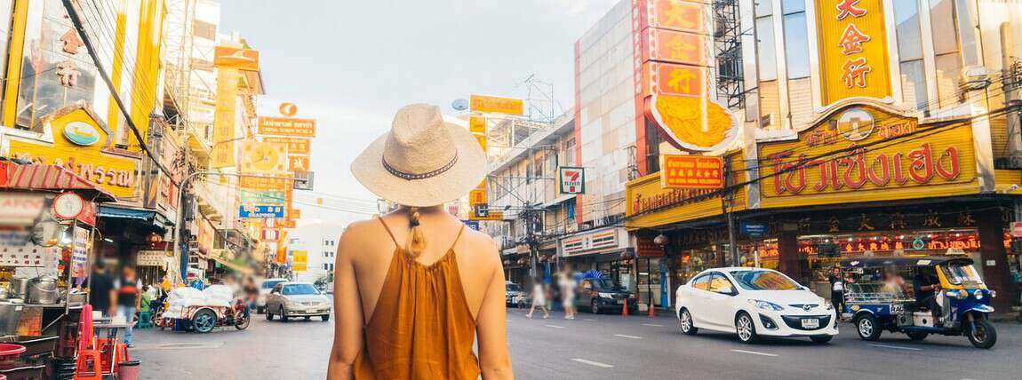 A woman wearing a brown dress and wide-brimmed hat walking along a bustling street lined with vibrant signs and shops in an Asian city.