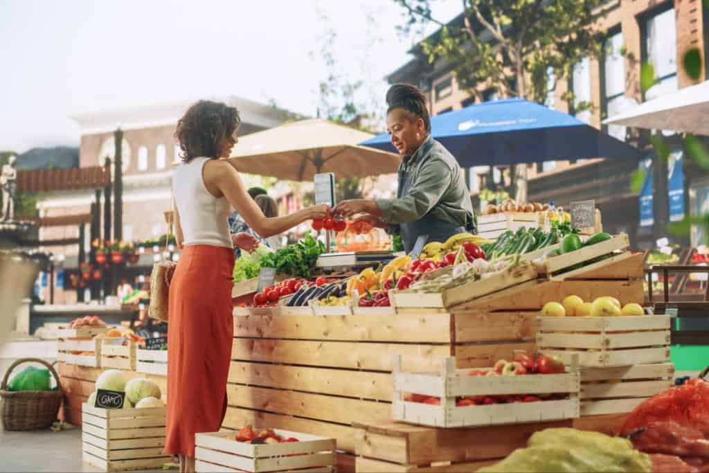 A woman shopping for fresh fruits and vegetables from a vendor at an outdoor farmers market. The vendor is handing her a bag of produce, with various fruits and vegetables displayed on wooden crates u.