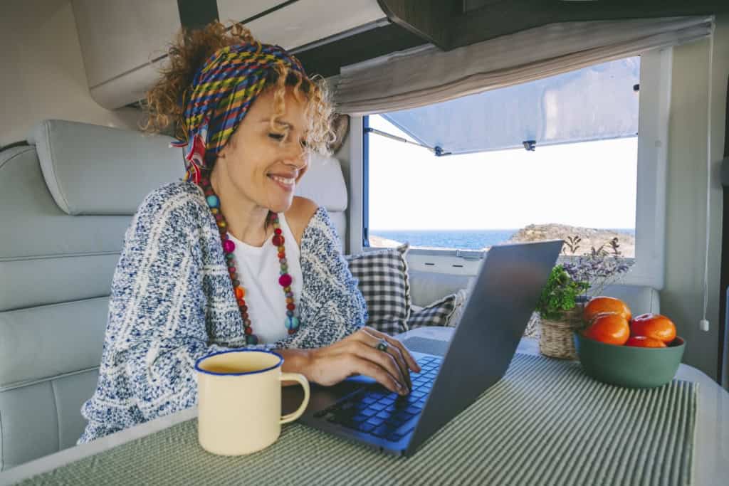 A female digital nomad with curly hair, colorful headband, and vibrant jewelry working on a laptop inside a camper van with a view of the ocean through the window, enjoy the expat life.