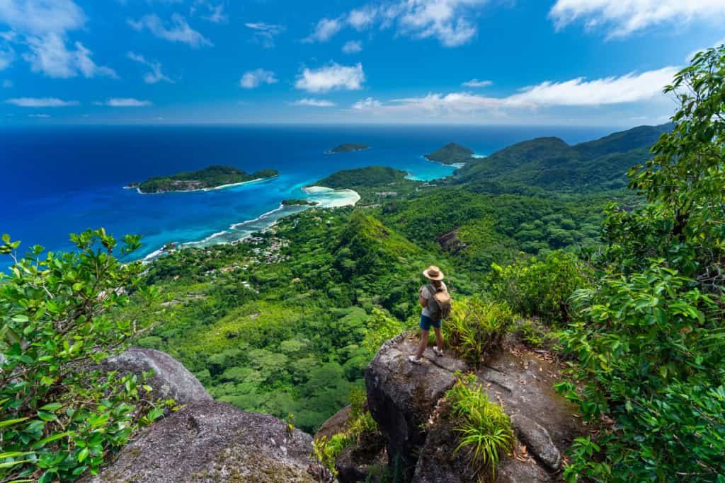 A hiker standing on a large rock overlooking a lush green tropical landscape with islands and the ocean in the background.