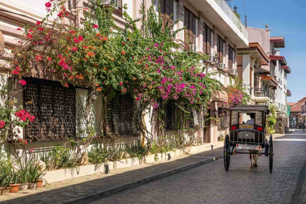 A charming European street scene featuring a cobblestone road, vibrant pink and red flowers cascading from balconies, and a vintage horse-drawn carriage traveling along the picturesque street.