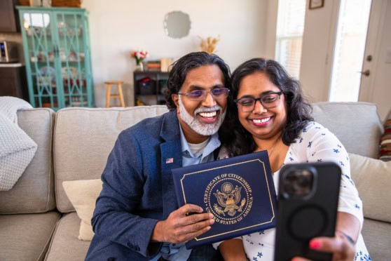 Smiling couple of diverse ethnicity taking a selfie with a US passport, representing international citizens securing health insurance coverage in the United States.