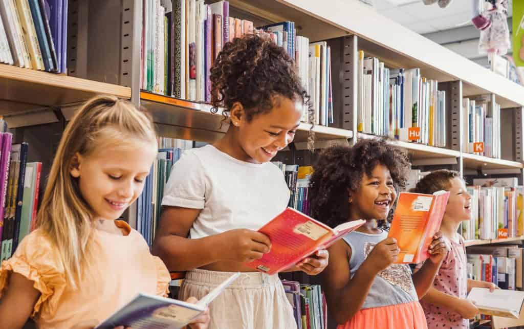 Group of diverse young international students reading books and smiling in a library aisle, promoting literacy and learning.