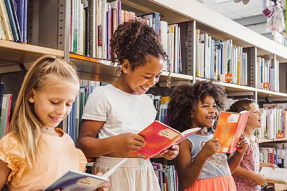 Group of diverse children reading books and smiling in a library aisle, promoting literacy and learning.