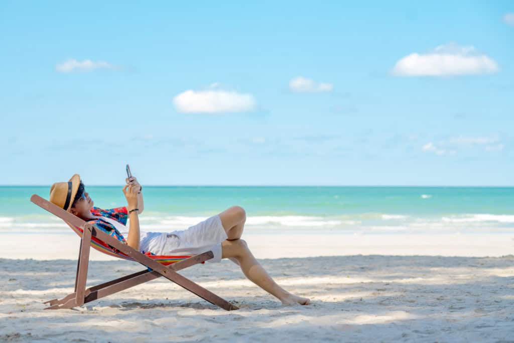 a male expat relaxing in a deck chair on an empty beach, using his cell phone to manage his finances abroad