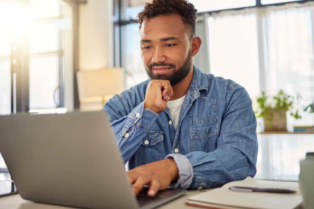 A young man with a beard and casual denim shirt studying abroad on his laptop in a well-lit office space with large windows and indoor plants.