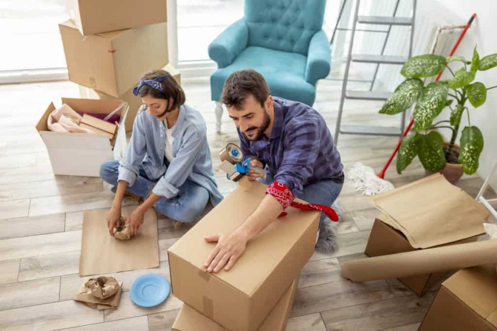 Young couple packing boxes before moving abroad in a bright, modern living room. They are surrounded by packing materials, including tape, paper, and boxes, preparing for their relocation.