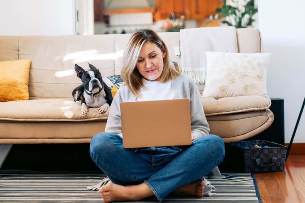 Woman sitting cross-legged on the floor with a laptop, smiling, in a cozy living room with a beige sofa and decorative pillows, researching moving abroad and jobs