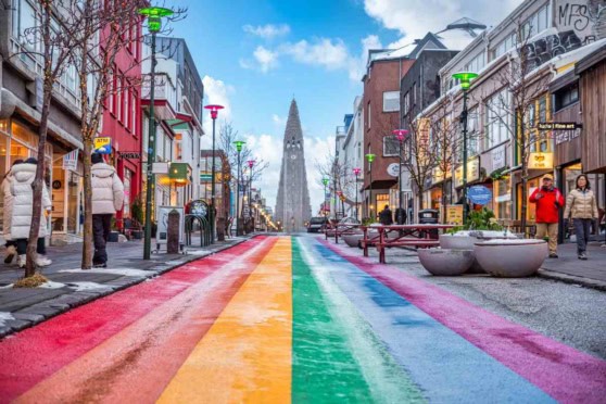 Vibrant rainbow-colored crosswalk stretching across a busy urban street with pedestrians, shops, and the iconic spire of a city landmark in the background.