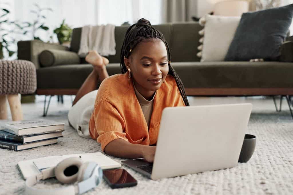 A young woman with braided hair, wearing an orange shirt, lying on her stomach on a cozy rug while working on her laptop, studying abroad in a modern living room.