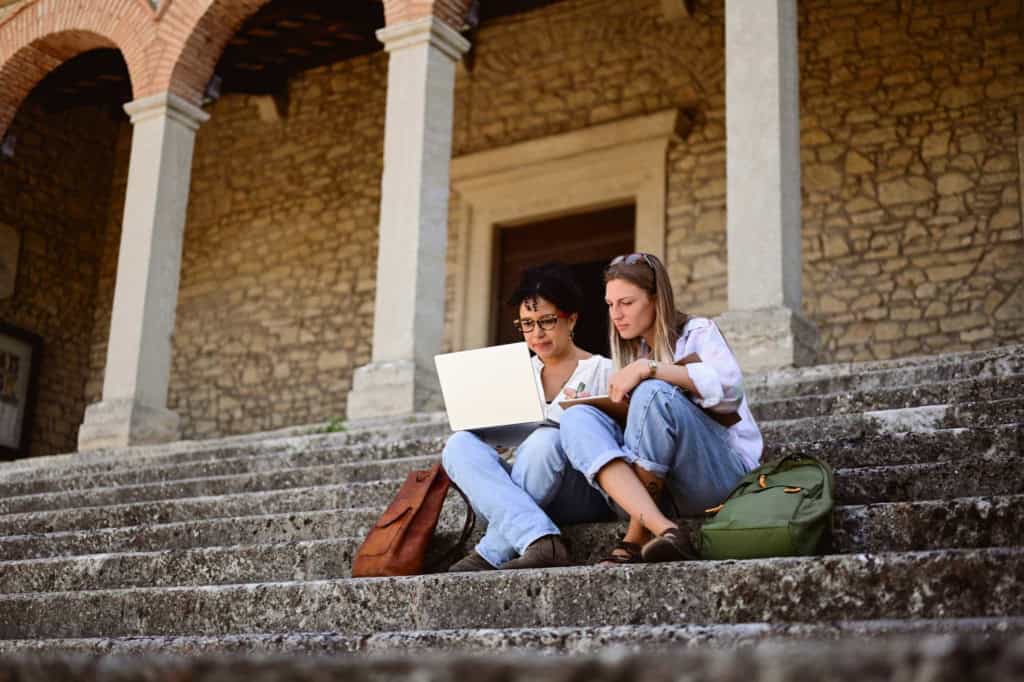 Two young women sitting on stone steps, studying abroad together with a laptop and notebooks, in front of a historic stone building with arches and columns.