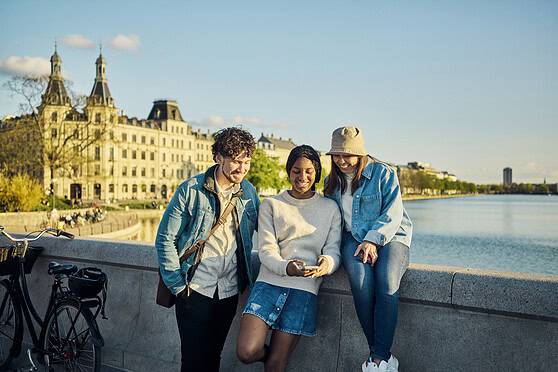 A diverse group of three young adults enjoying a sunny day by the river, with historic buildings in the background. They are casually dressed, smiling, and looking at a smartphone, capturing a moment.