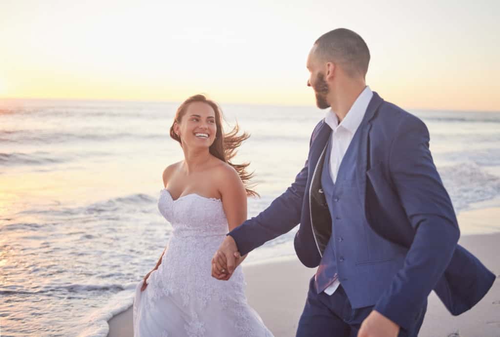 Wedding couple walking hand in hand along the shoreline at sunset, getting married abroad, enjoying a romantic moment on the beach. The bride is smiling and wearing a white wedding dress, while the groom is dressed in a blue.
