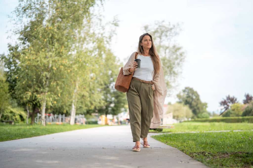 a woman looking relaxed and walking through a park, holding a cup og coffee, taking a break from remote work