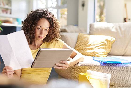 Woman with curly hair reading a document on a tablet in a bright living room, with plants and cushions, during her international health insurance research.