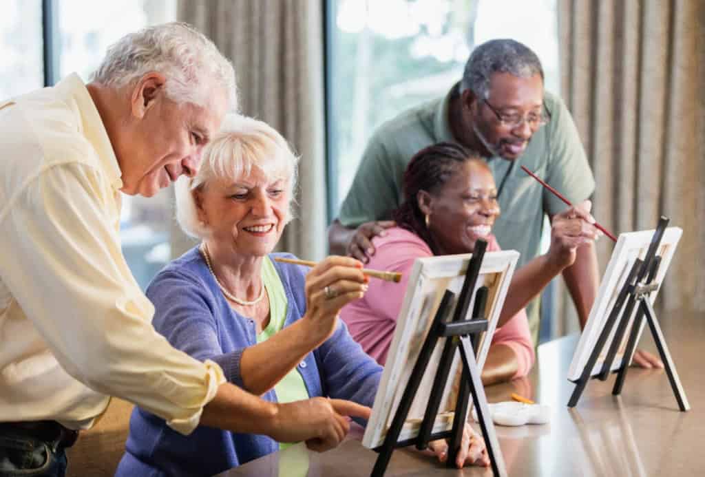 Elderly diverse group painting on canvases in a bright room, with a window showing greenery, during a retirement activity.