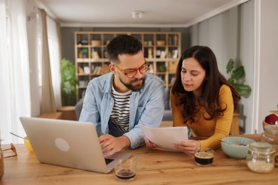 Couple reviewing documents at home with a laptop, in a well-lit living room, related to international travel or insurance topics.