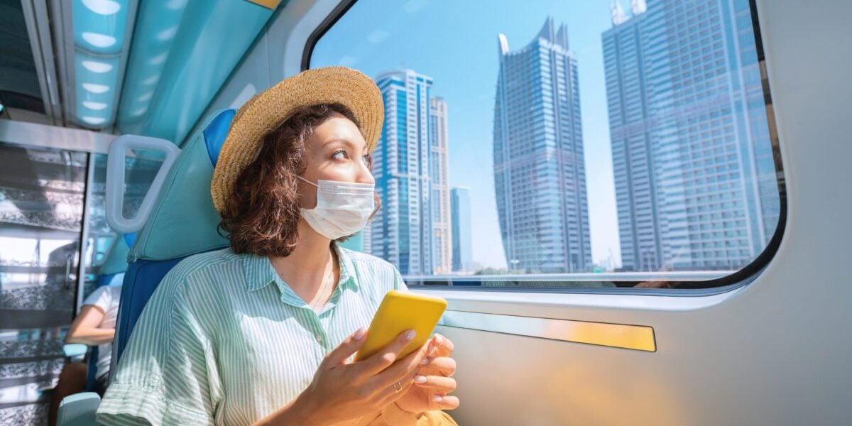 Woman in a protective medical mask rides the subway, holds a smartphone in her hands and looks out the window where the giant skyscrapers of Dubai rise Woman in a protective medical mask rides the subway, holds a smartphone in her hands and looks out the window where the giant skyscrapers of Dubai rise