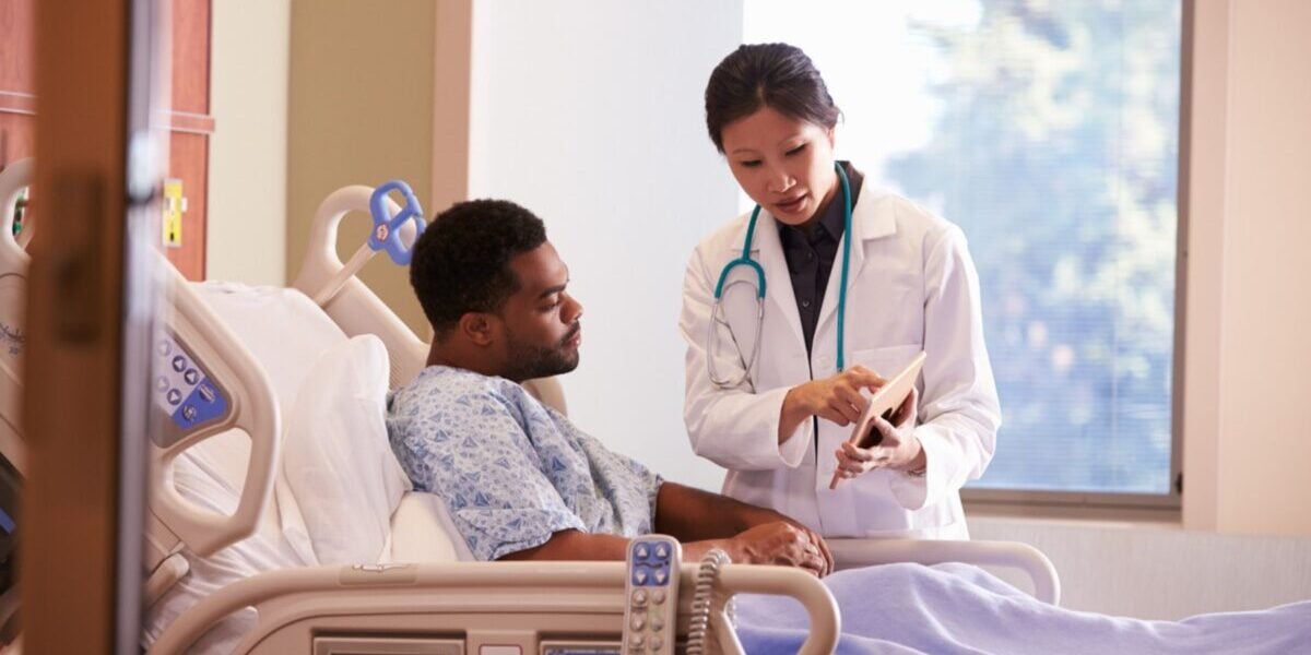 hospital-patient-in-bed a male patient laying in a hospital bed talking to a female doctor, reflecting on the importance of annual health insurance reviews for ensuring adequate hospital coverage